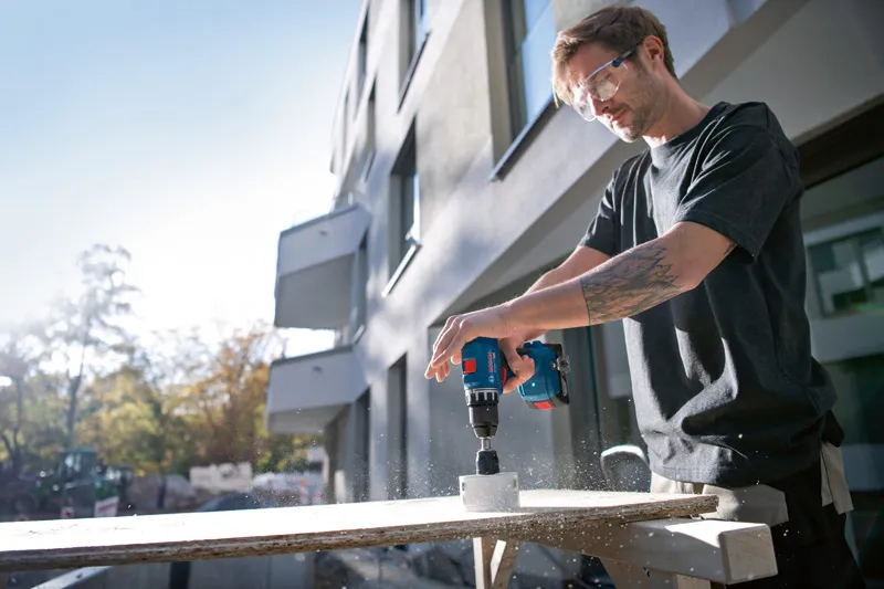 En person iført sikkerhedsudstyr borer et hul i træ udendørs med en boremaskine.